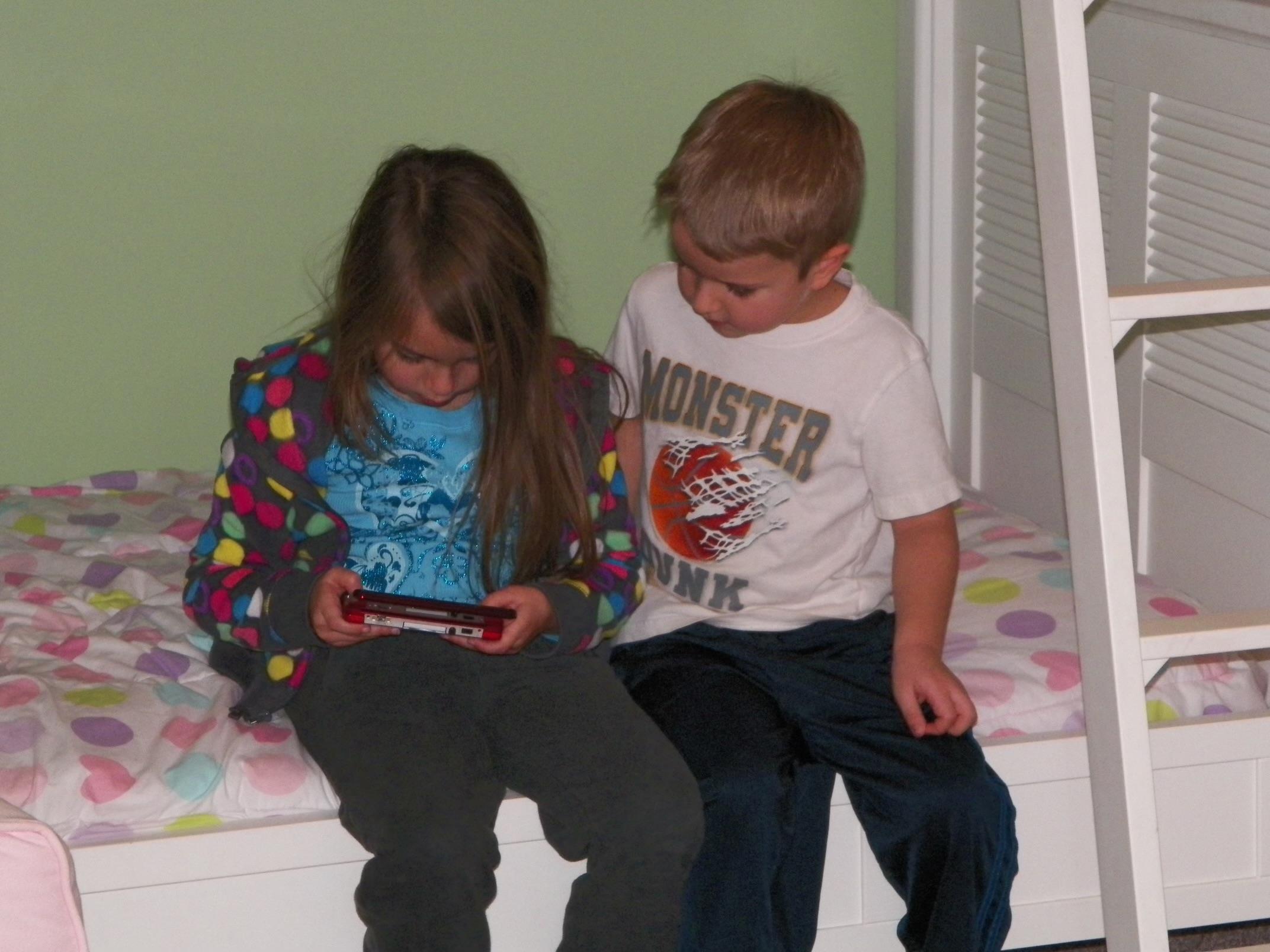 Two young children share a moment playing on a device in a bright, playful bedroom.