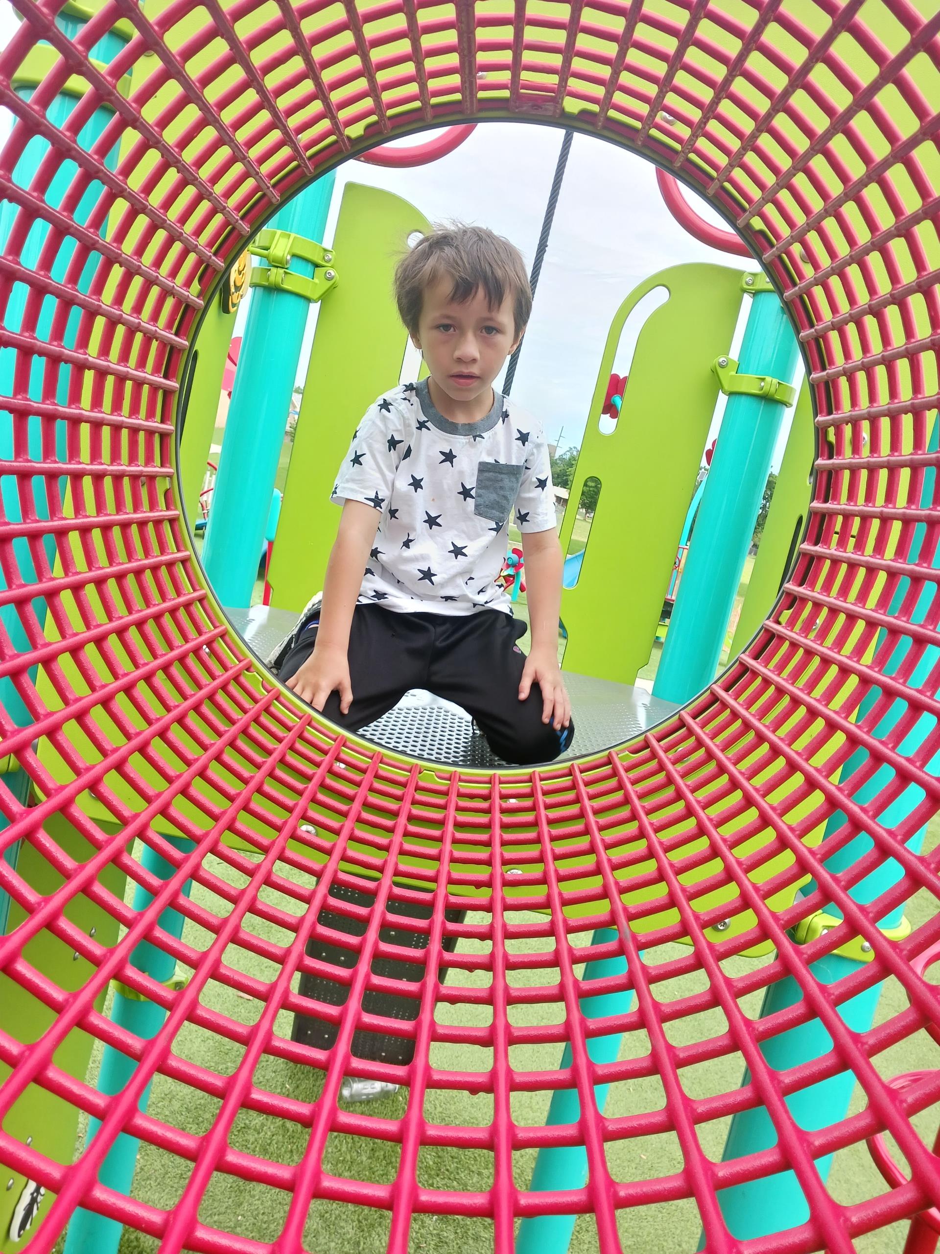 Young boy with a serious expression sitting on a playground piece, viewed through a colorful tube.