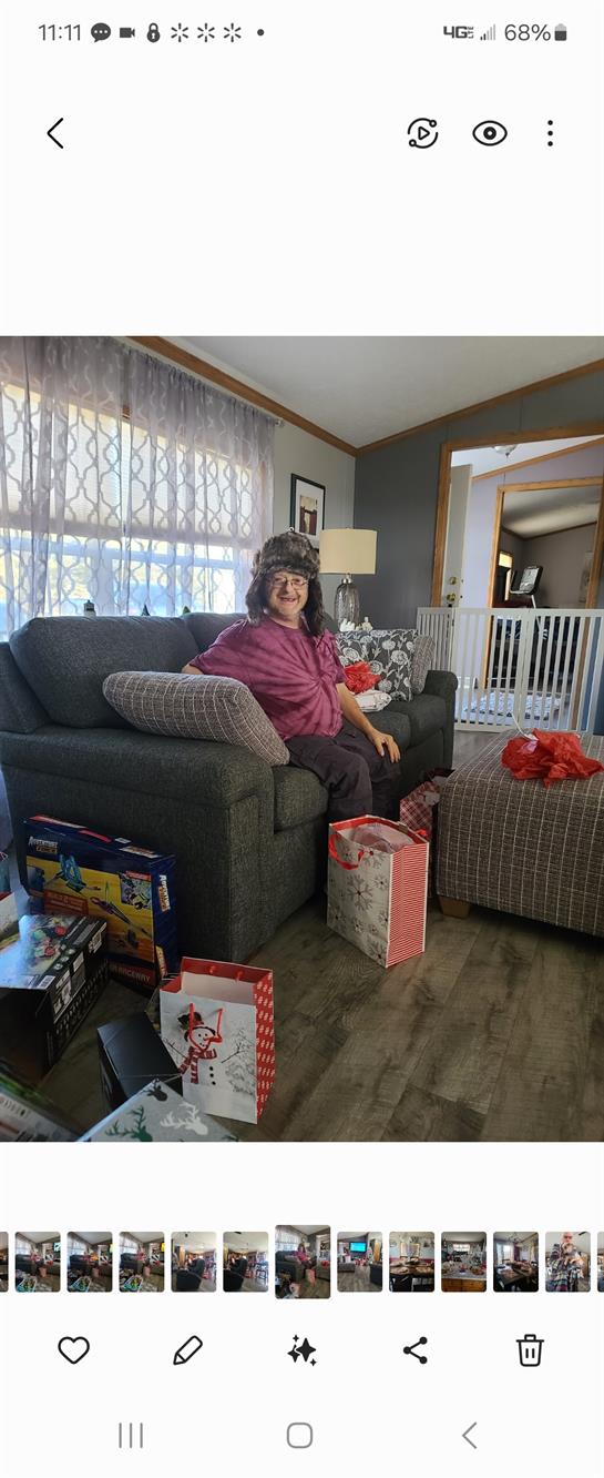 A woman sits comfortably on a couch in a living room with gifts around her. She wears a hat.