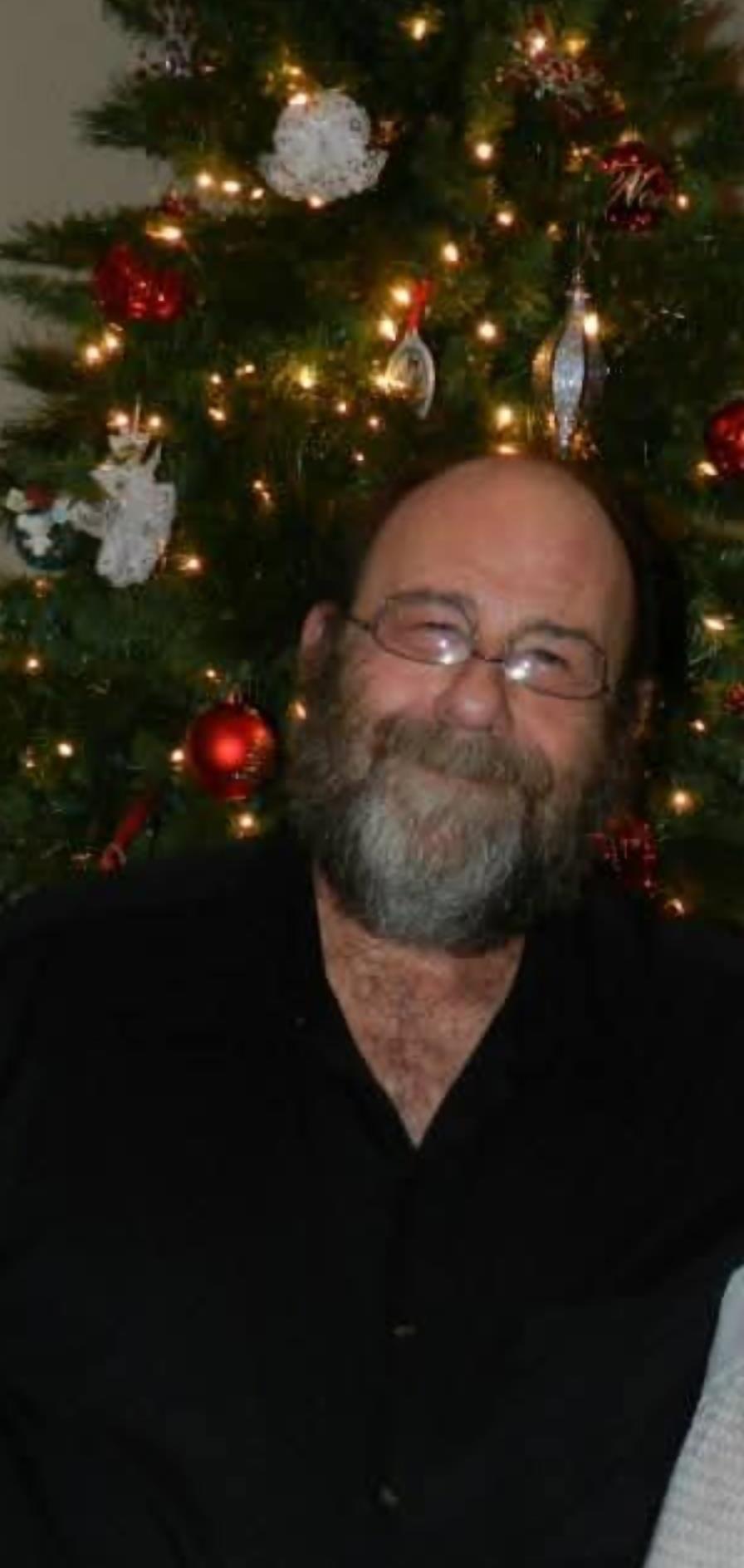 A smiling man with glasses and a beard sits by a beautifully decorated Christmas tree.