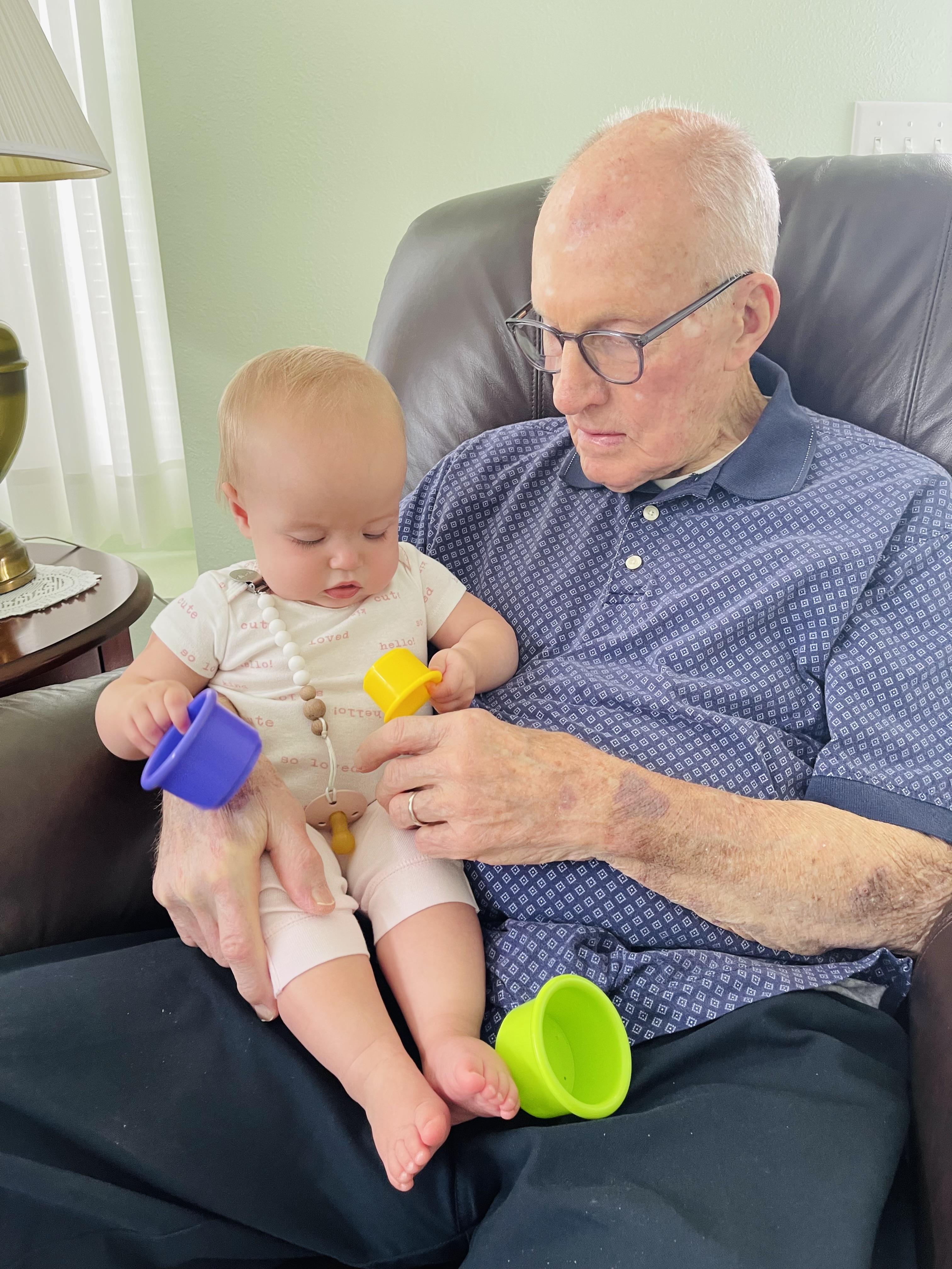 Elderly man and baby engaged in playful activity with colorful cups in a comfortable chair.