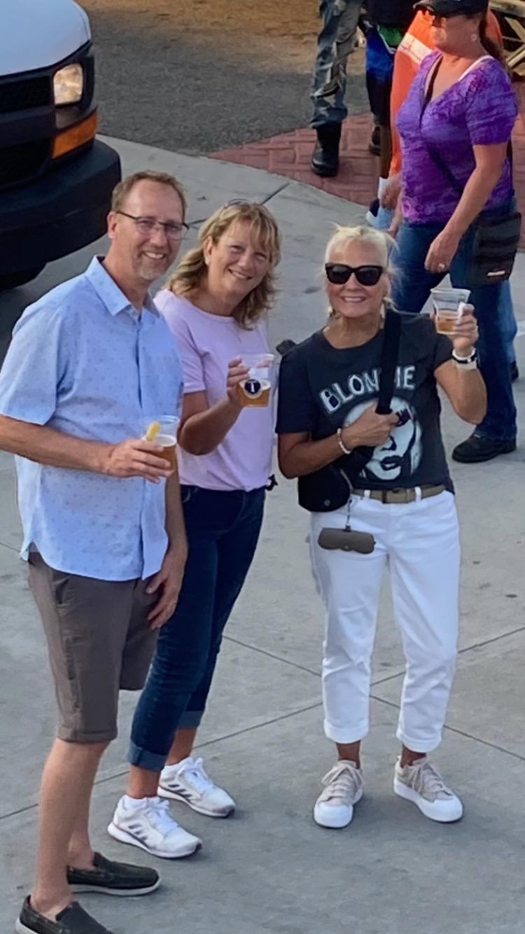 Three friends smile and pose with drinks, enjoying a sunny afternoon in a lively park atmosphere.