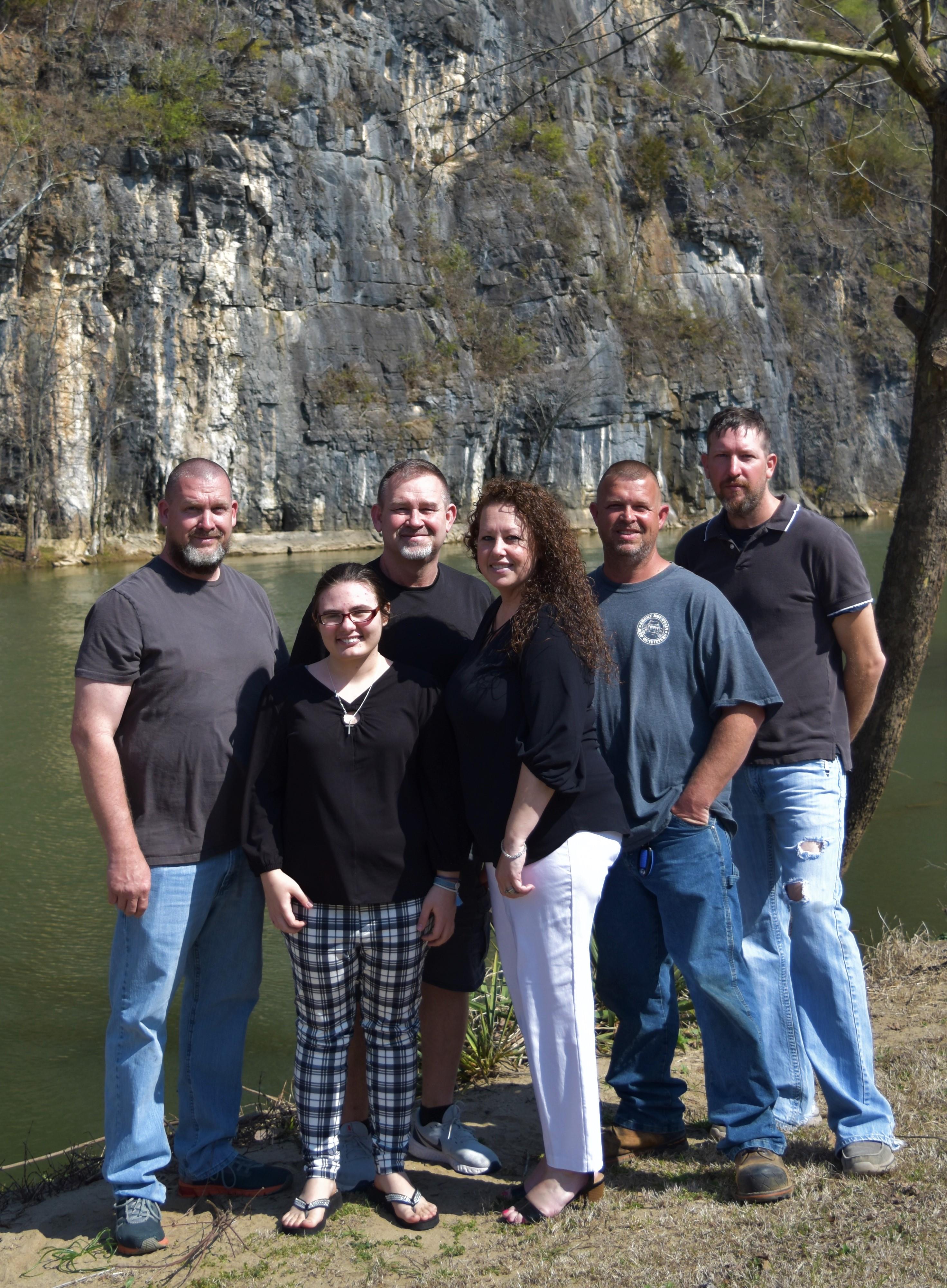 Five adults and one child pose joyfully by a calm lake with rocky cliffs behind them.