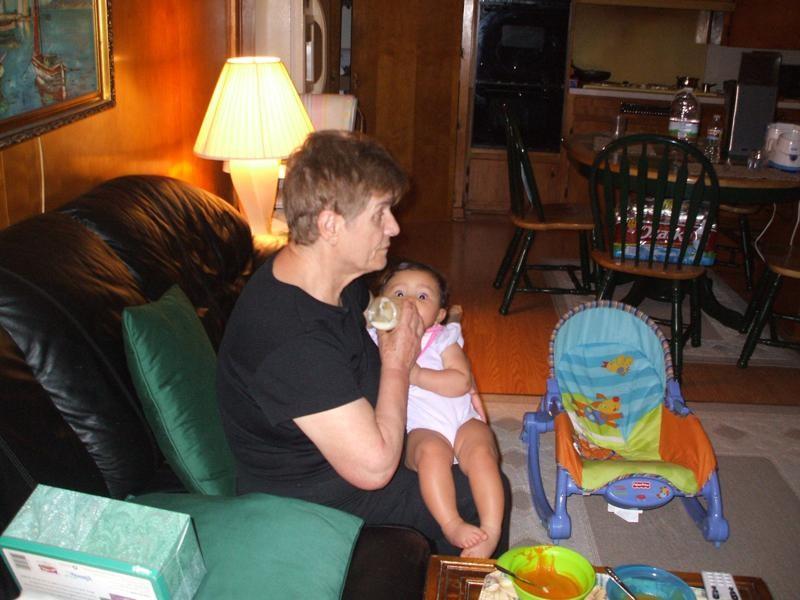 A grandmother feeds her grandchild in a cozy, furnished living room.