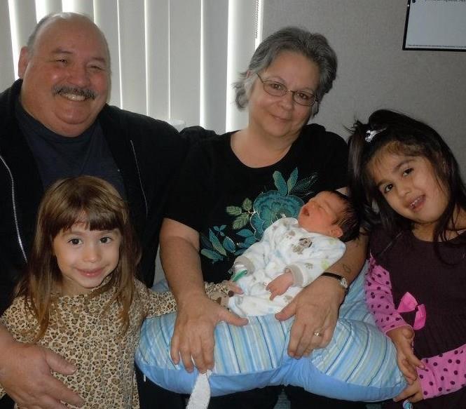 Children and grandparents joyfully hold a newborn baby, enjoying a warm family moment indoors.