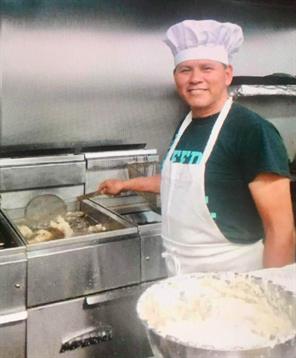 Chef skillfully cooks fried dishes while wearing a hat and apron in a bustling kitchen.
