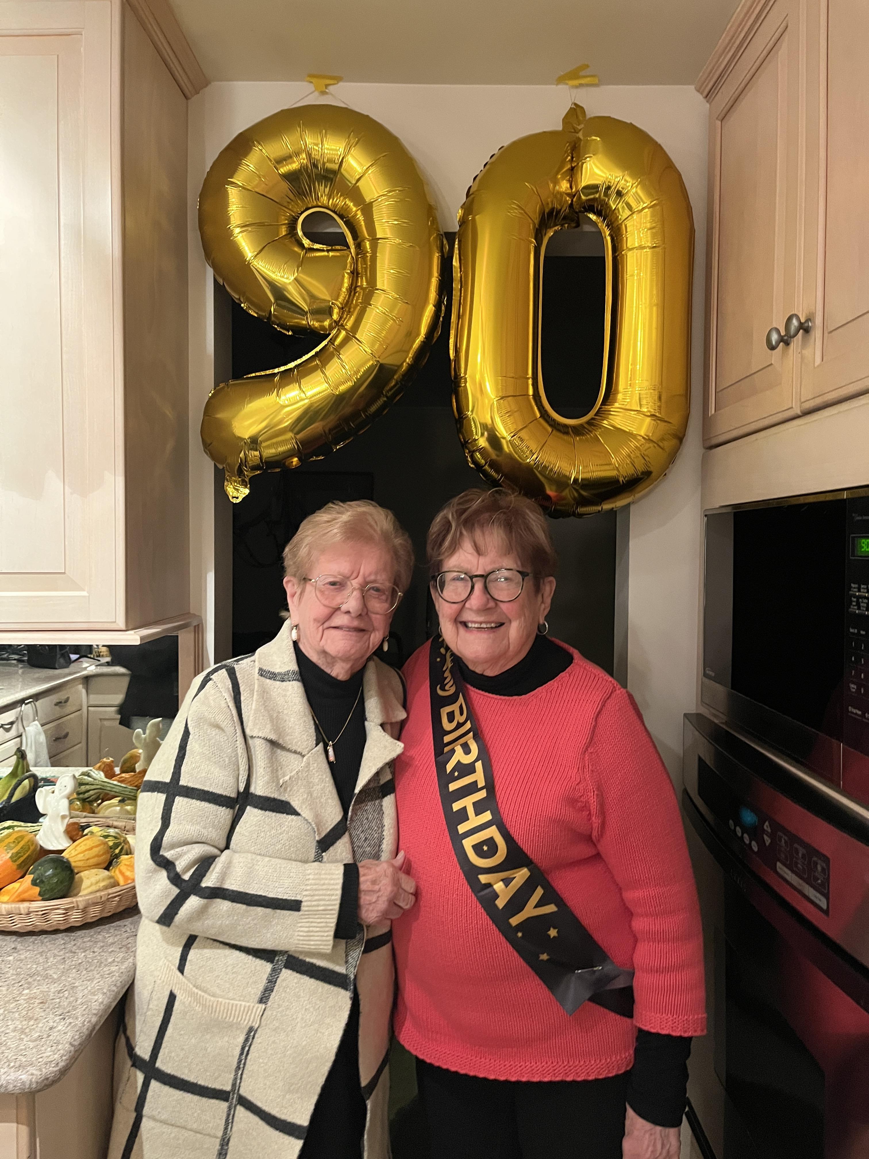 Two women celebrate a 90th birthday together in a festive, balloon-decorated kitchen.