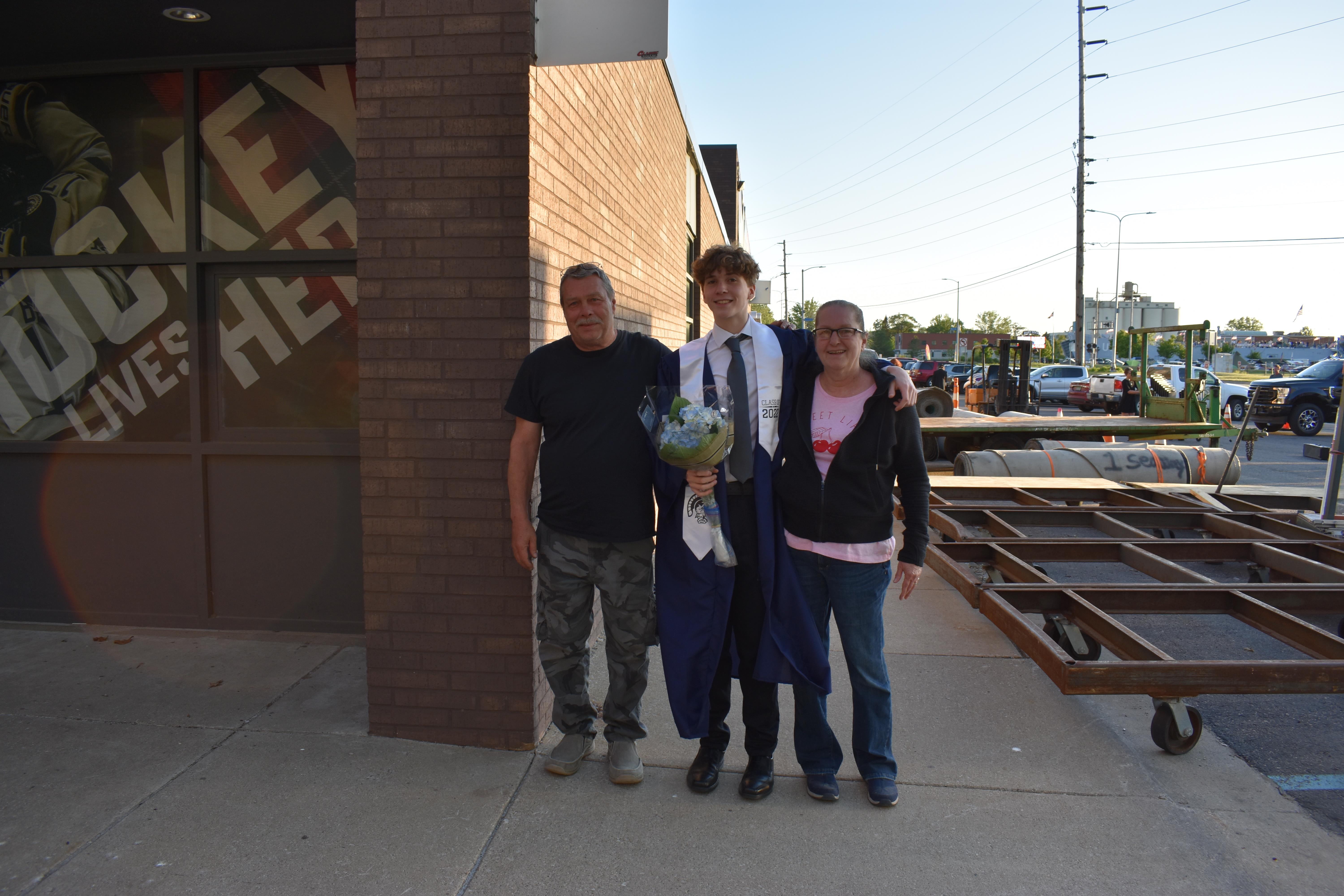 A family group stands outside a building, smiling while holding a small dog, enjoying a sunny day.