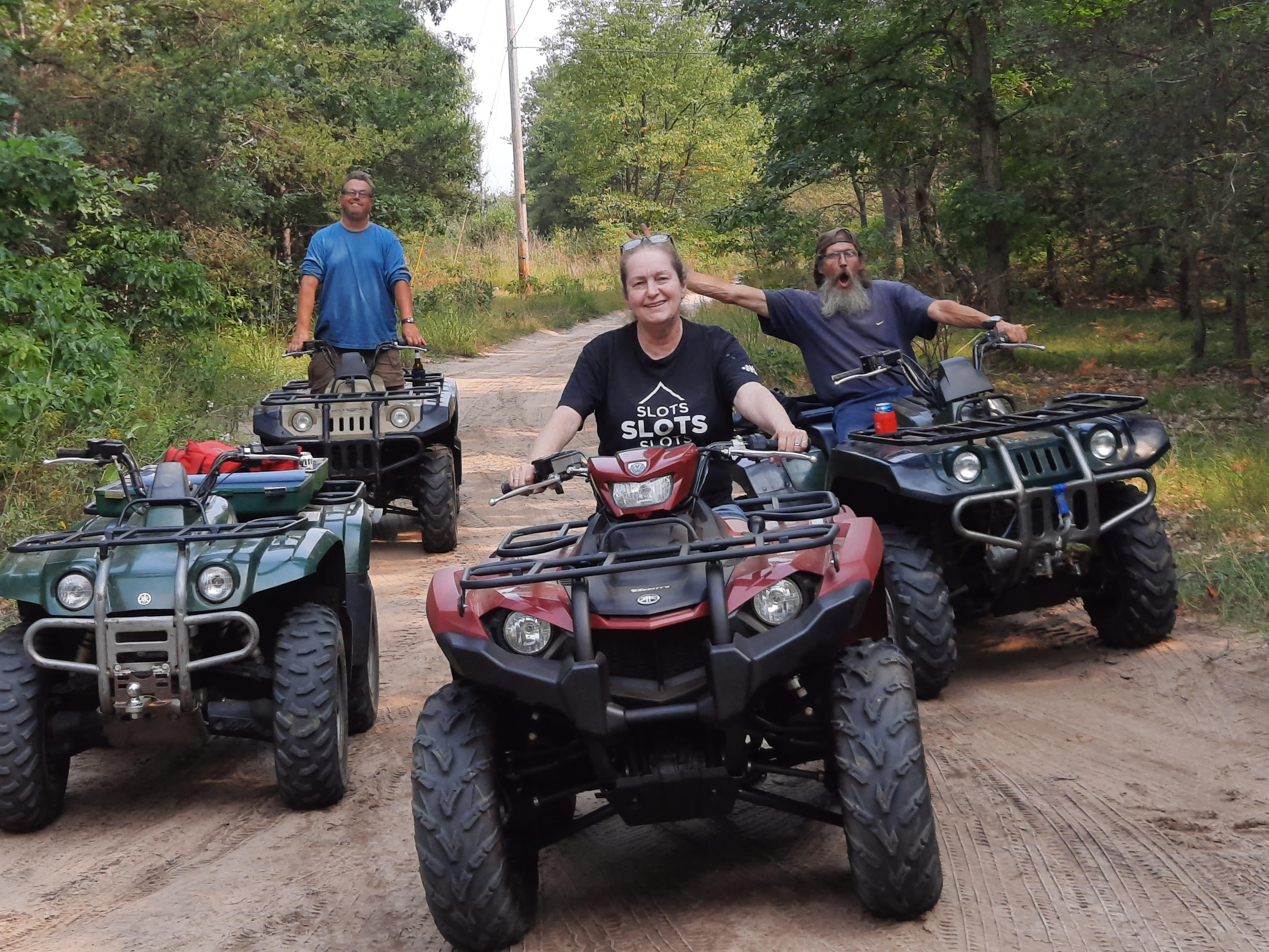 Friends ride all-terrain vehicles on a dirt path surrounded by trees during a sunny day.