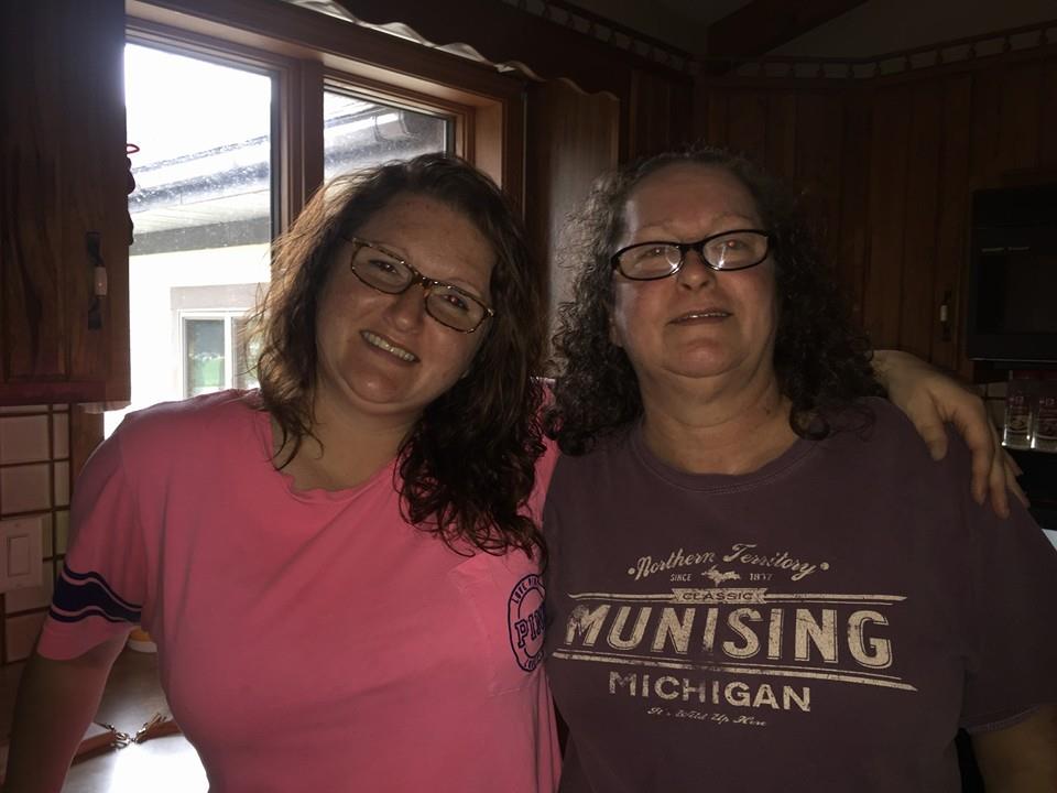 Two women pose with smiles in a warm kitchen, enjoying a joyful moment together.