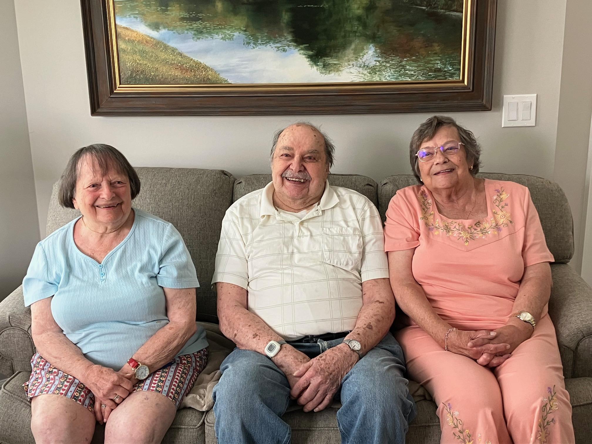 Three elderly people smile together on a couch in the living room.