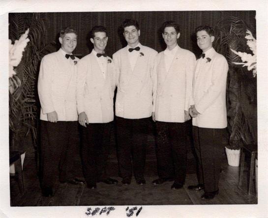 Five young men dressed in tuxedos with bow ties stand together, smiling warmly.