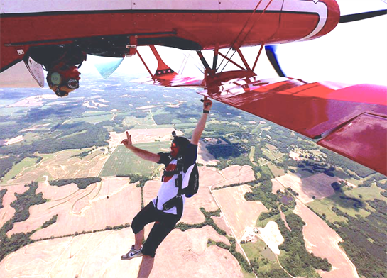 A person hangs from the wing of a biplane while soaring high over beautiful rural fields.