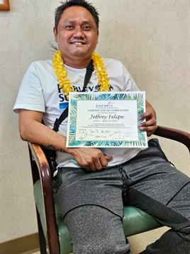 Individual proudly holds a certificate while wearing a lei, seated in a relaxed environment.