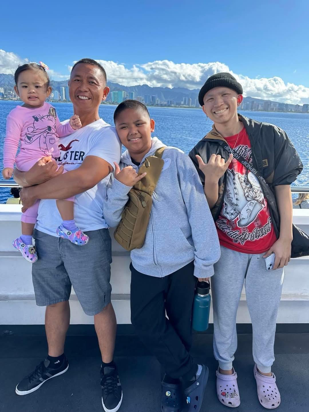 Four family members smile and pose together by the water under a clear blue sky.