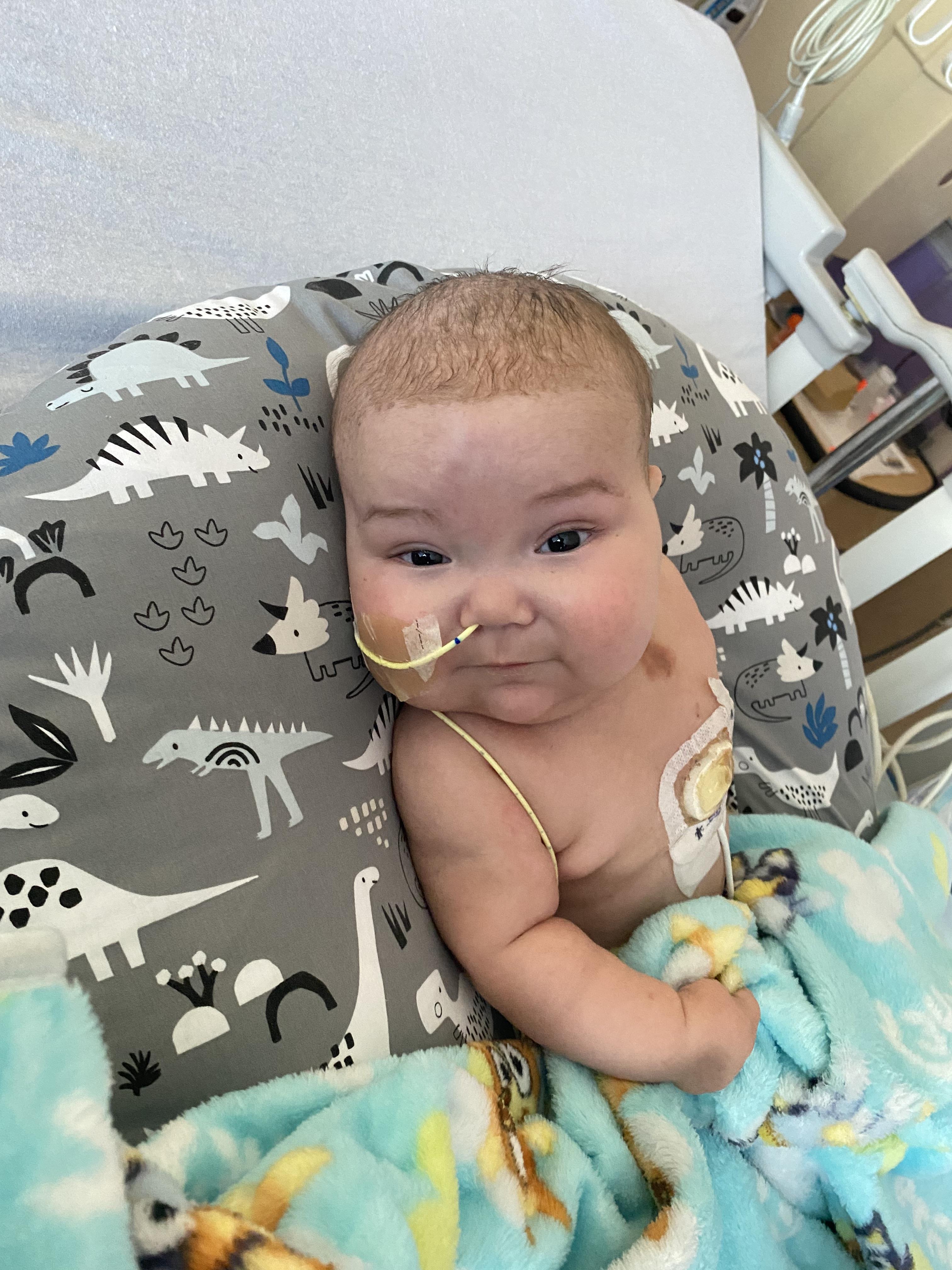 Young infant comfortably rests in a hospital bed with medical apparatus, showing resilient spirit.