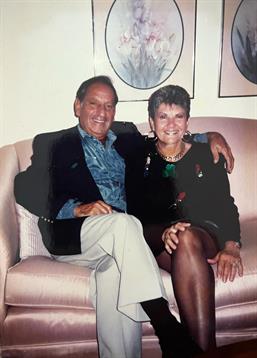 An elderly couple sits closely together on a comfortable sofa, smiling happily in a living room.