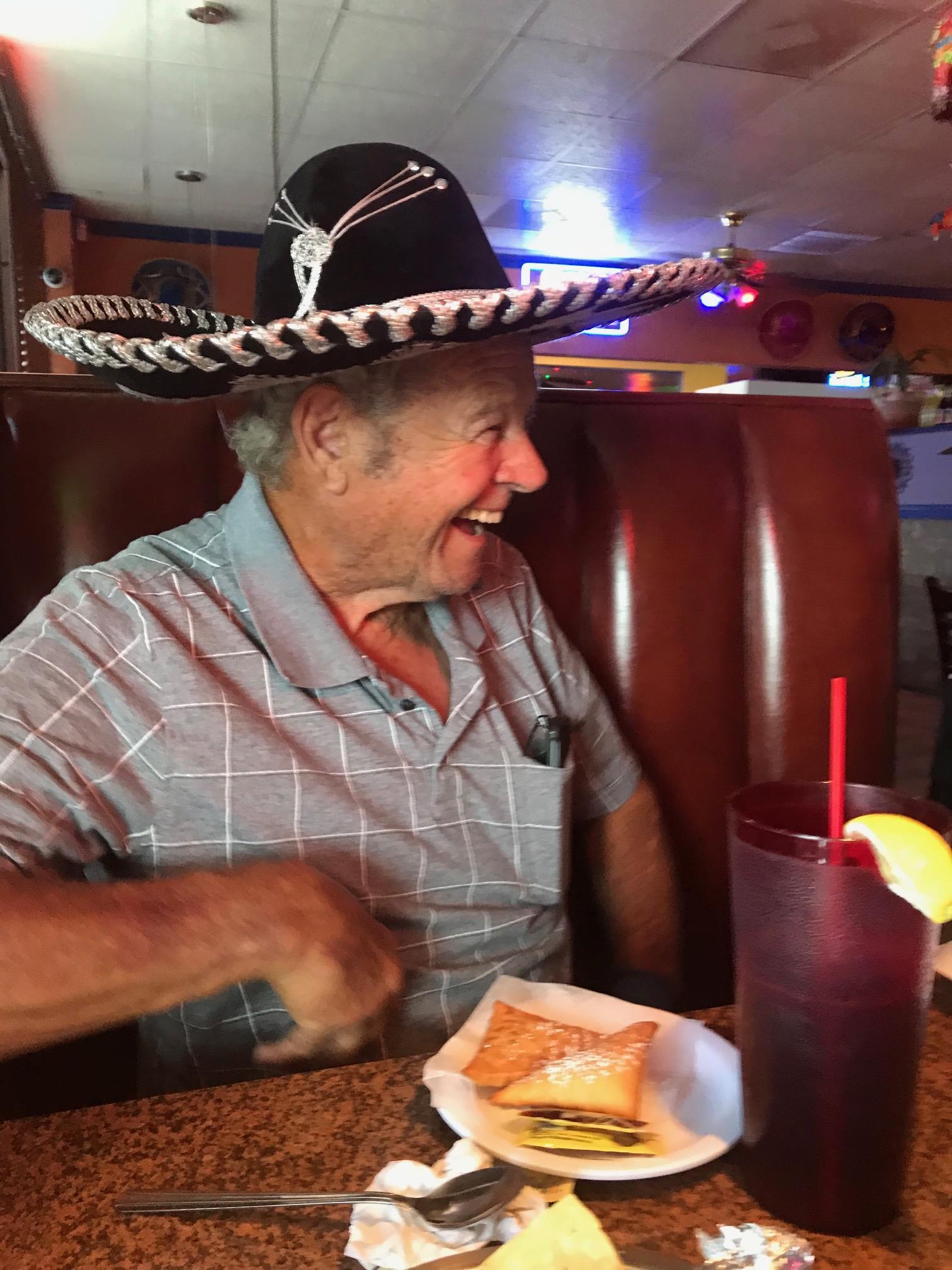 An elderly man smiles joyfully while seated in a restaurant, enjoying a meal and refreshing drink.