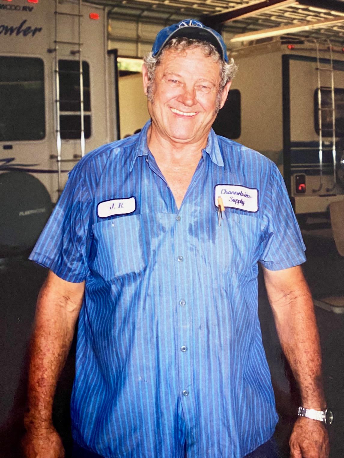 A cheerful mechanic stands in a repair shop, wearing a blue uniform and smiling broadly.
