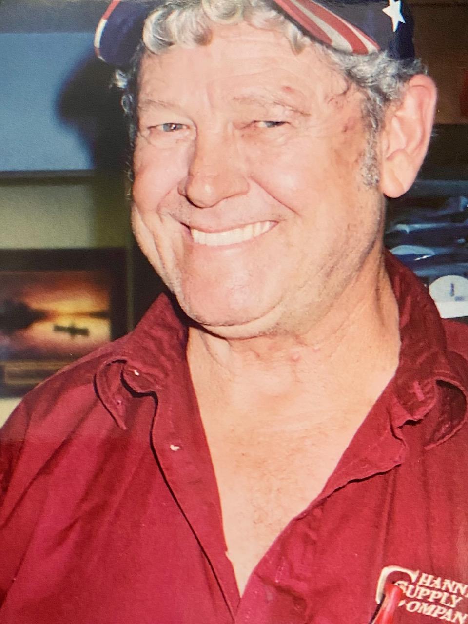 A cheerful elderly man in a red shirt and cap smiles warmly in a decorated indoor space.