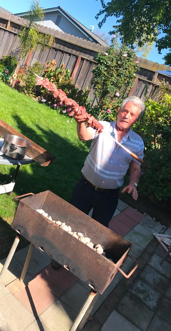 A man skillfully holds a large skewer of meat over a grill in a vibrant backyard setting.