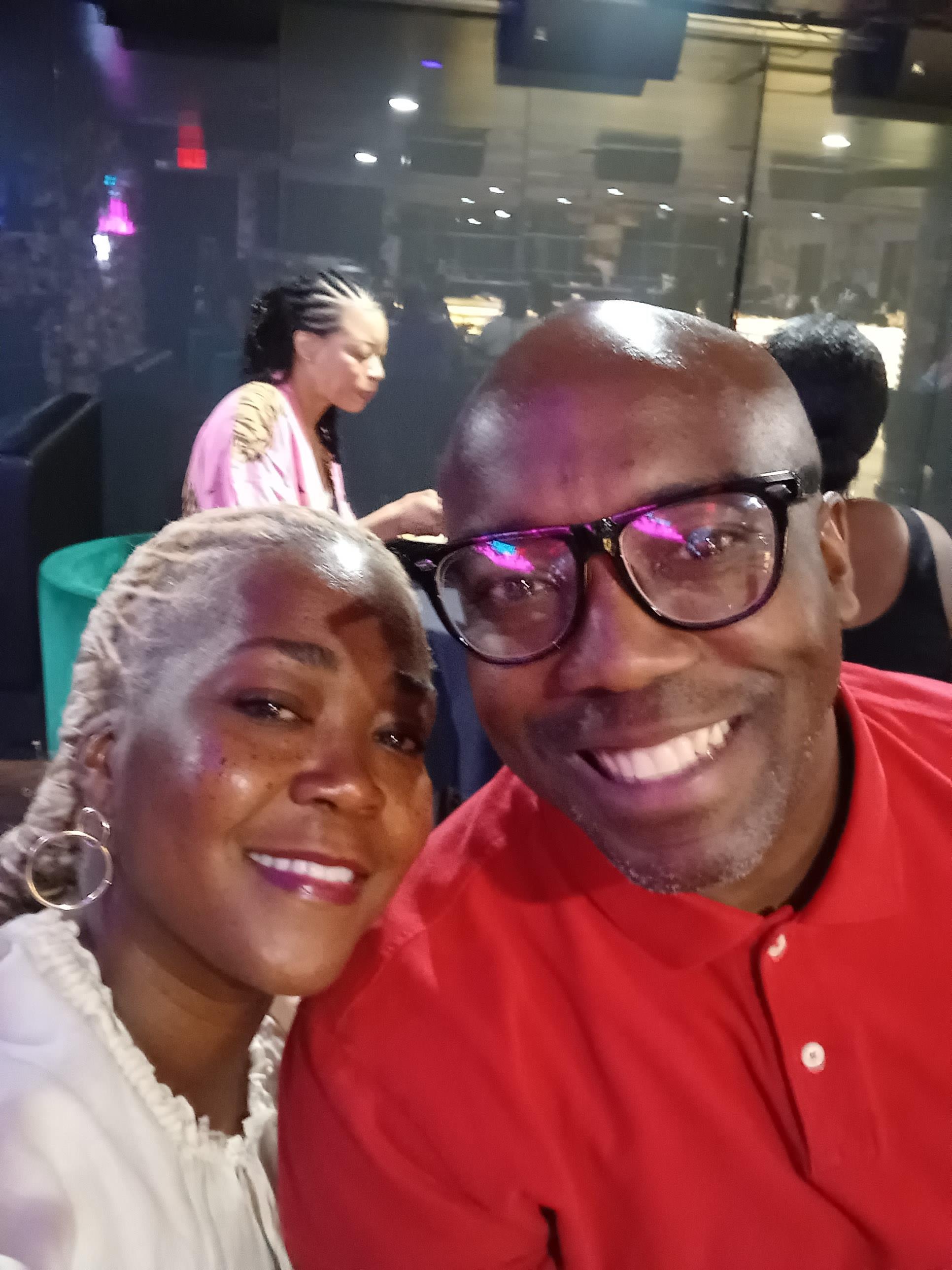 A joyful couple poses for a selfie amidst a lively atmosphere with friends around.