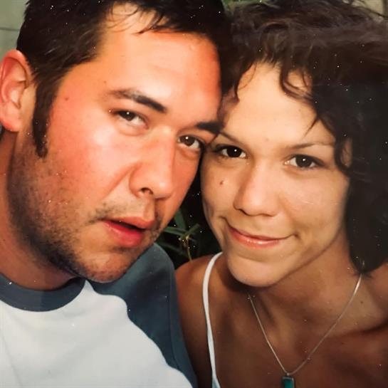 A couple poses closely together, smiling happily in a casual outdoor setting on a warm day.