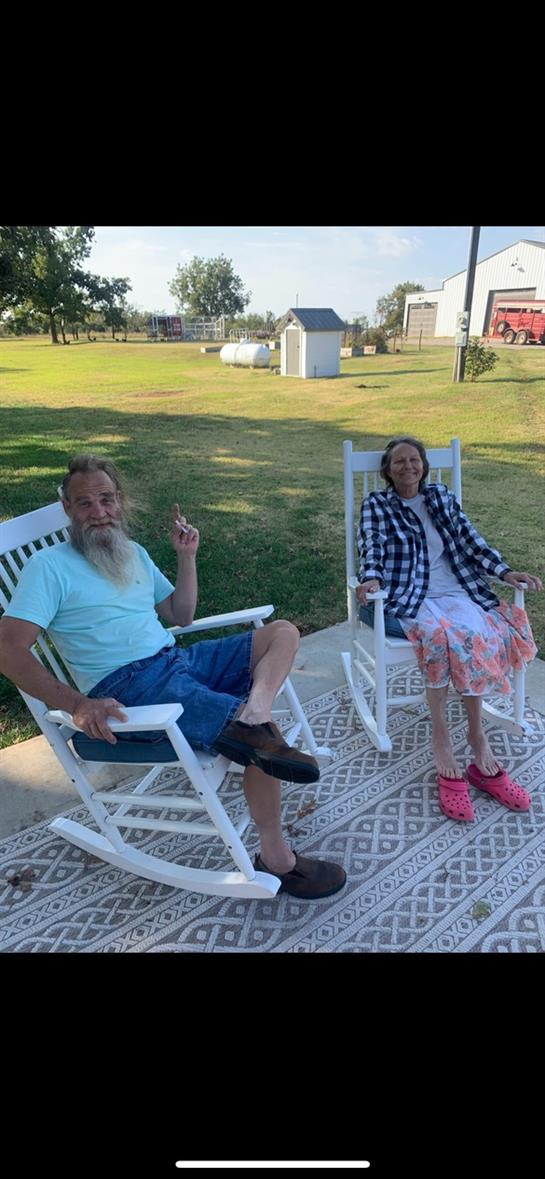 Older man and woman relax in rocking chairs, enjoying a sunny day in their backyard garden.