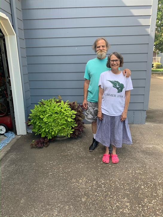 A couple smiles together outside their house, wearing casual clothing on a warm day.