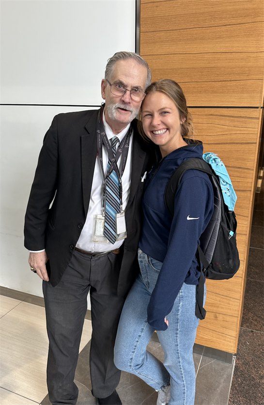 An older man in a suit poses with a young woman in casual attire, both smiling happily together.