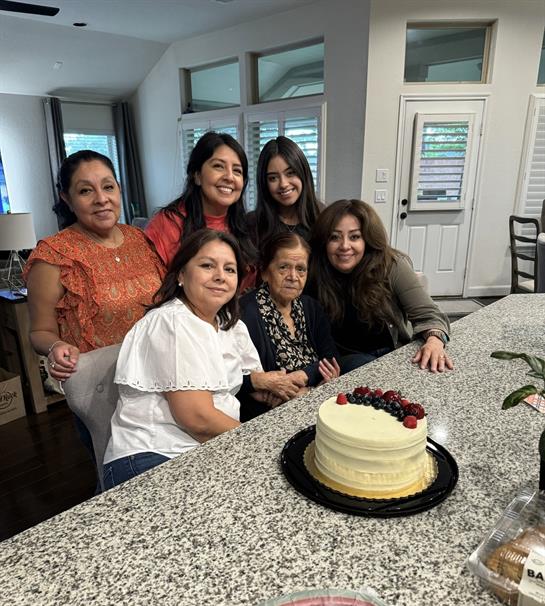 Six women celebrate together in a warm kitchen while enjoying a cake, showcasing laughter and hugs.