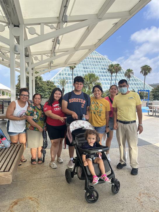 A group of eight enjoys leisure time outdoors, posing next to a stroller under a clear blue sky.