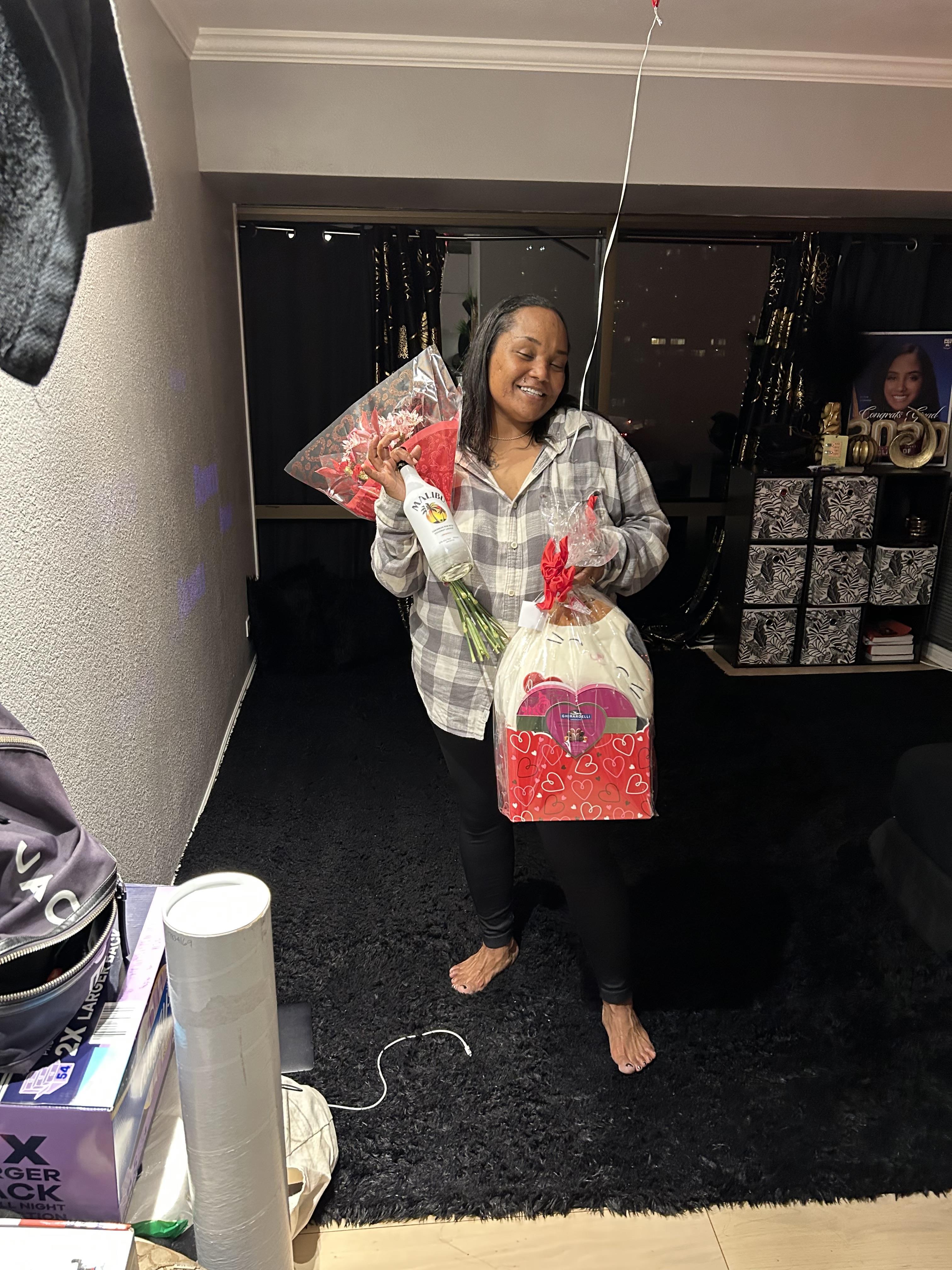 A woman joyfully holds wrapped gifts and drinks in a stylish living room during evening hours.