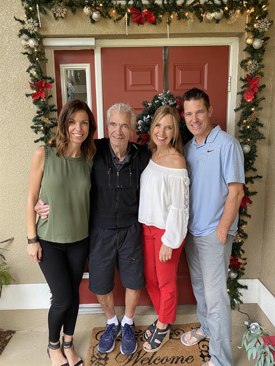 A group of four adults poses cheerfully at a holiday-decorated front door with greenery.