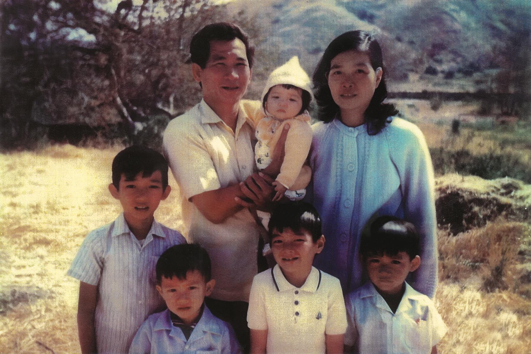 A family poses together outdoors in a sunny area, featuring six members across different ages.