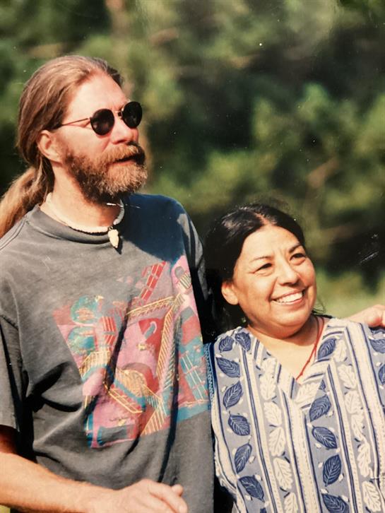 Two individuals are smiling together outside, enjoying each other's company in nature.