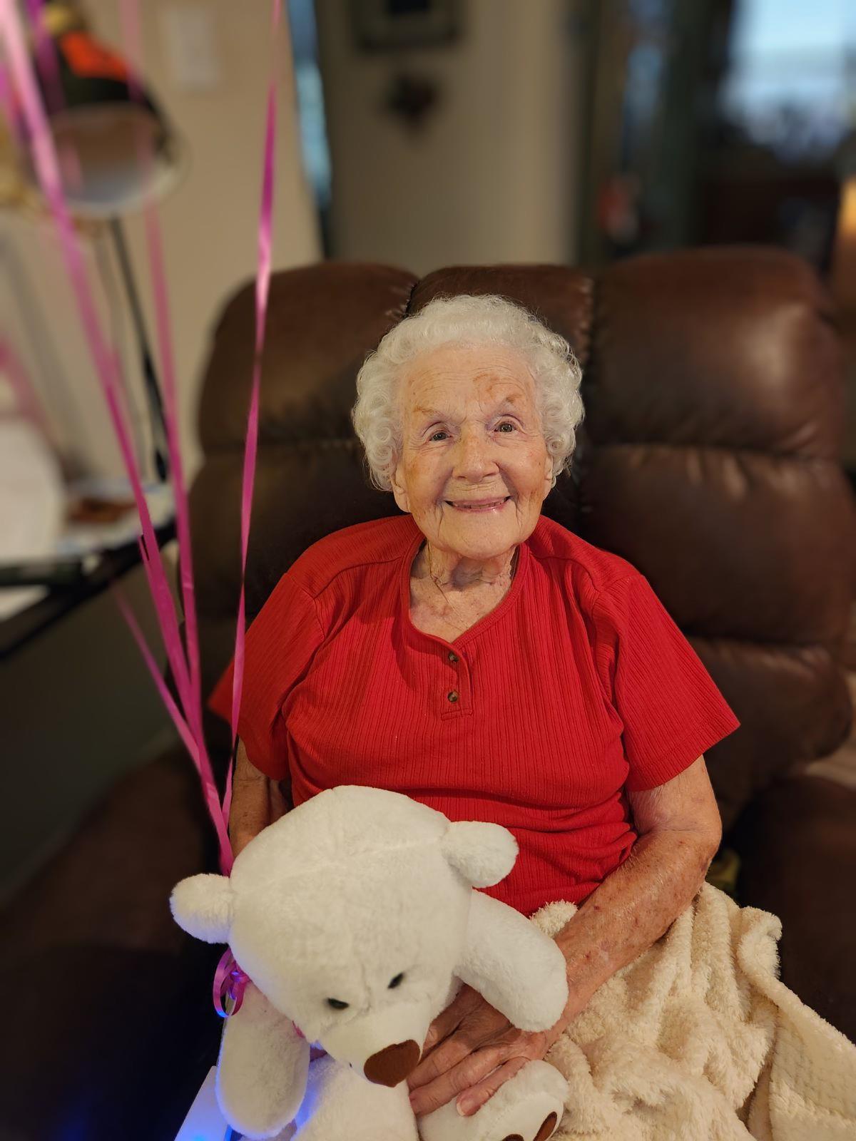An elderly woman sits comfortably in a chair, smiling with balloons and a teddy bear nearby.