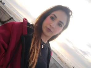 A woman stands by the beach, smiling warmly as the sun sets behind her, casting soft light.