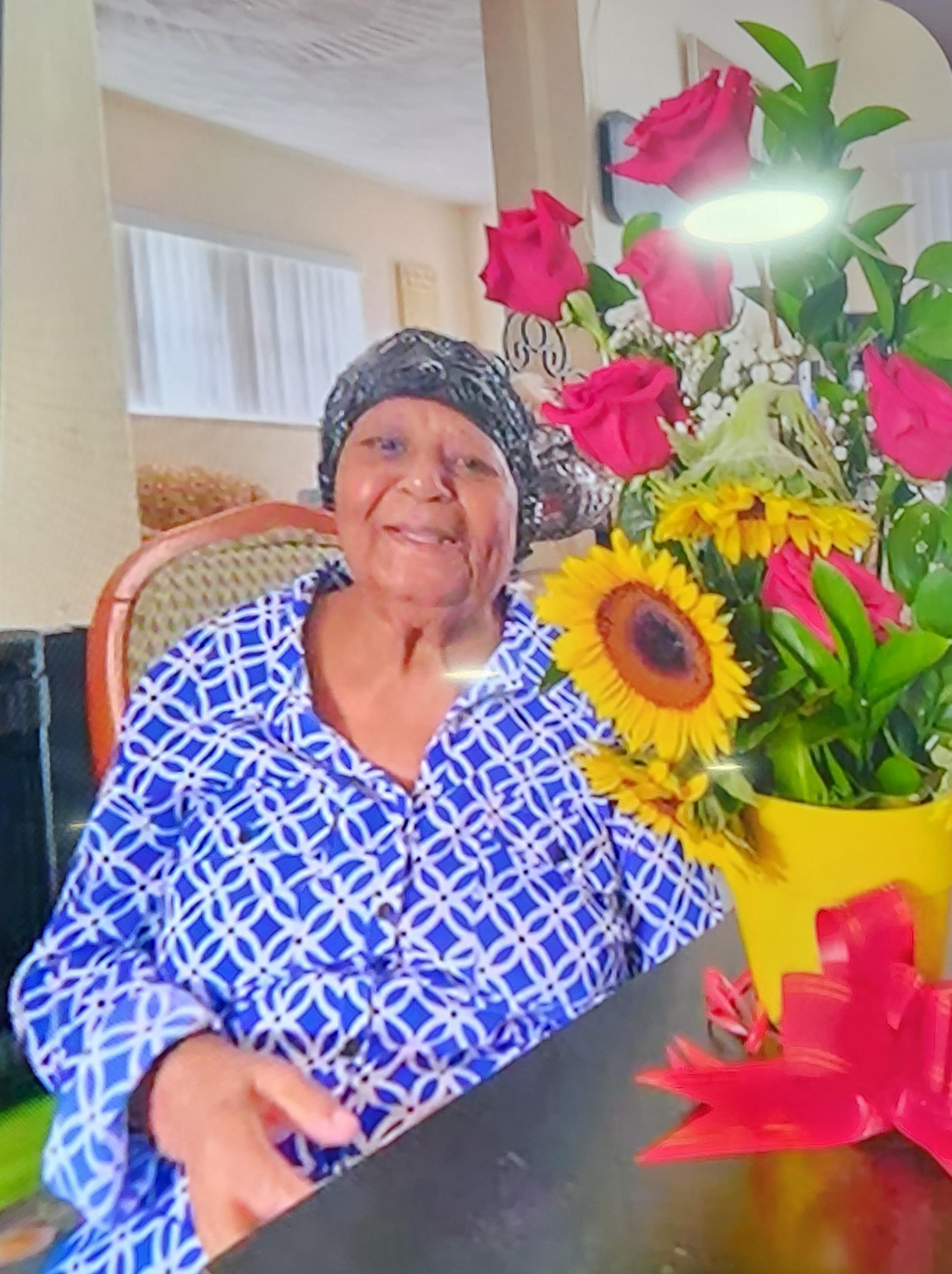An elder woman smiles brightly at a table adorned with vibrant flowers in a cozy space.
