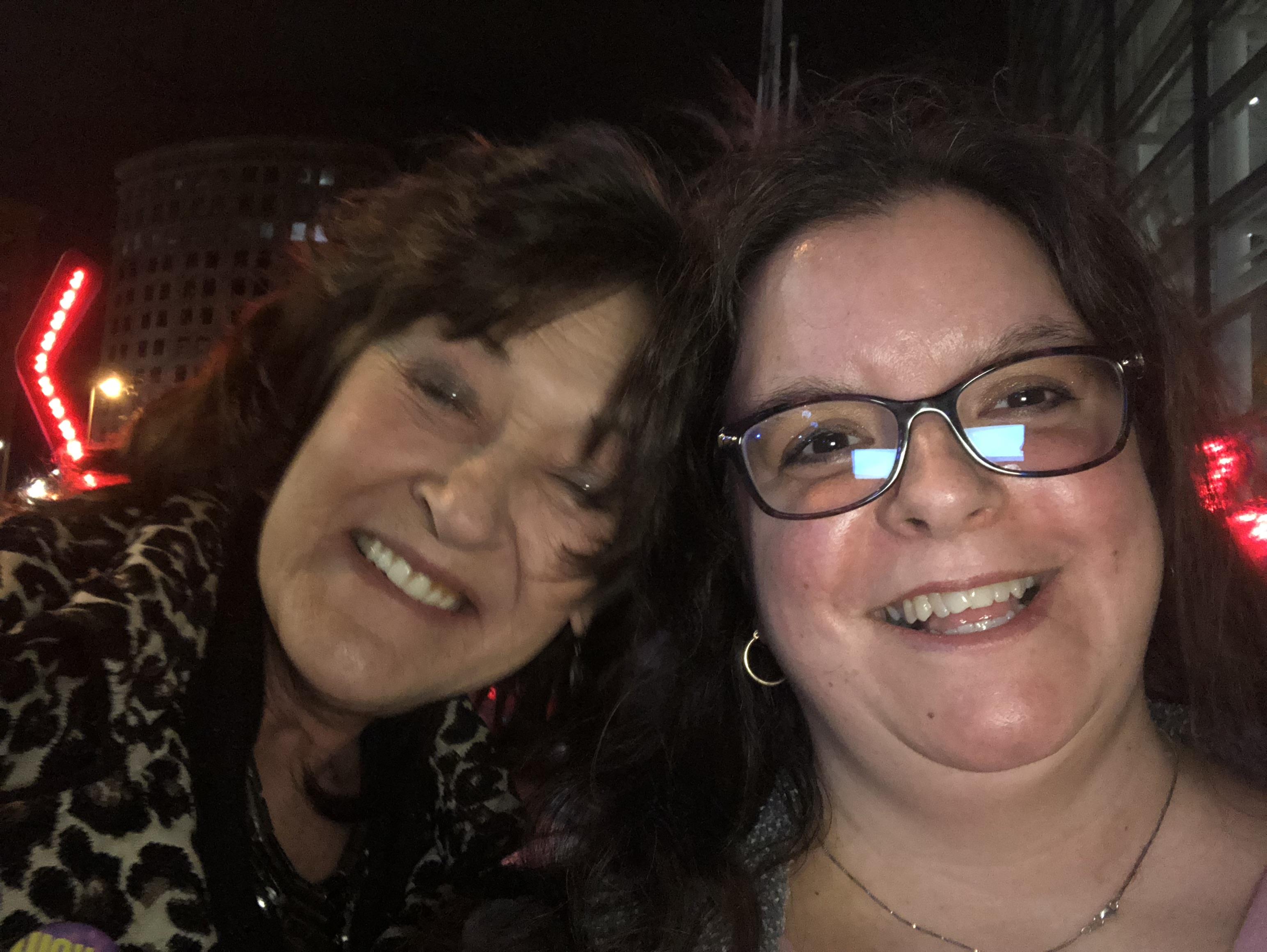 Two women are happily posing outdoors at night, surrounded by colorful lights and city vibes.