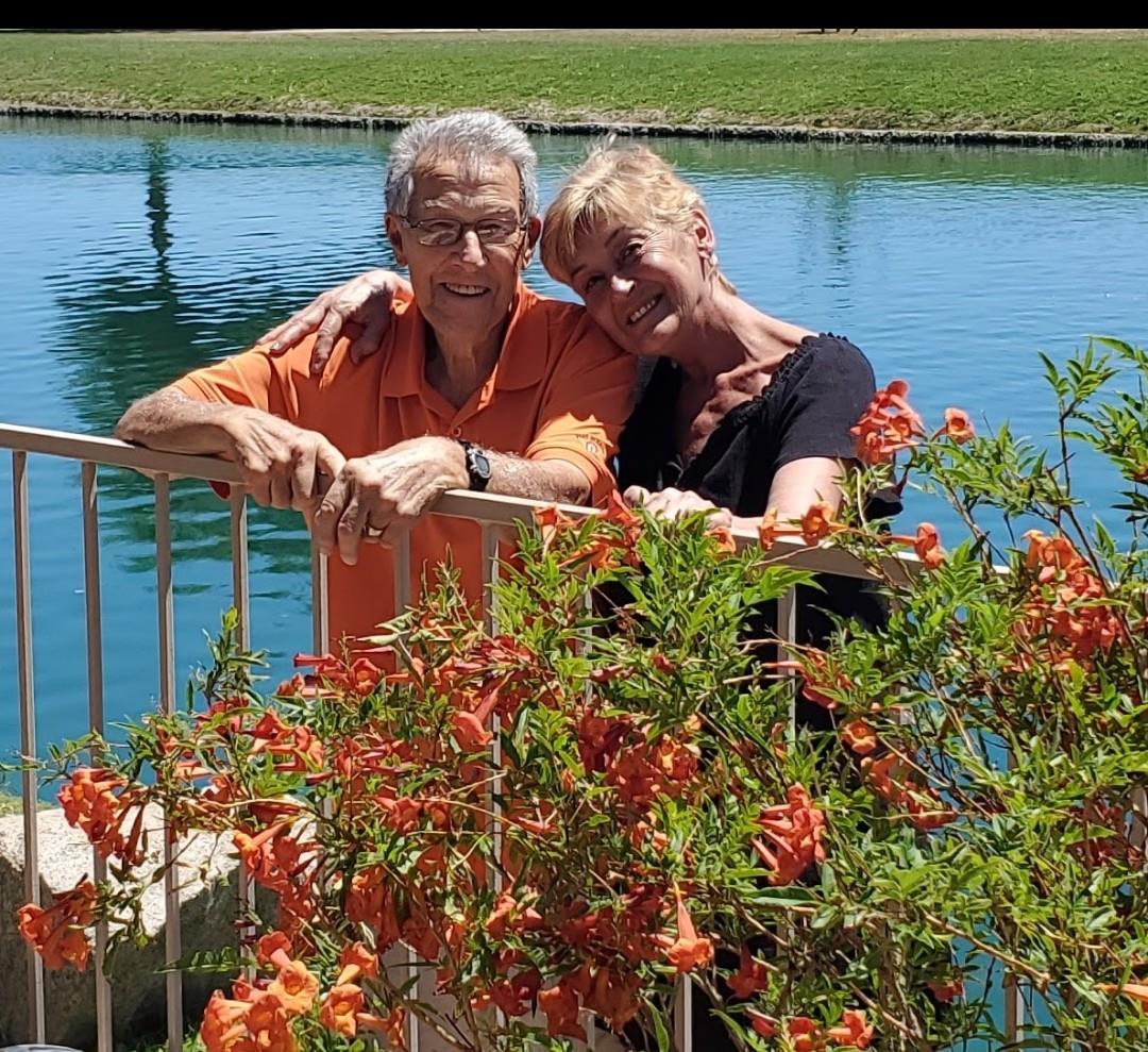 Two smiling individuals pose together by a scenic water body surrounded by blooming flowers.