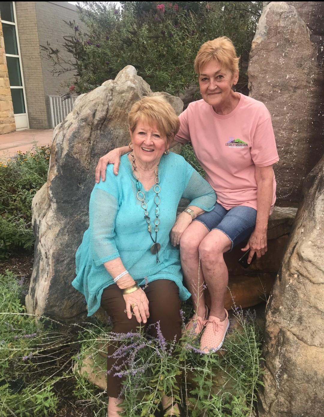 Two women smile together on large rocks surrounded by greenery in a garden setting.
