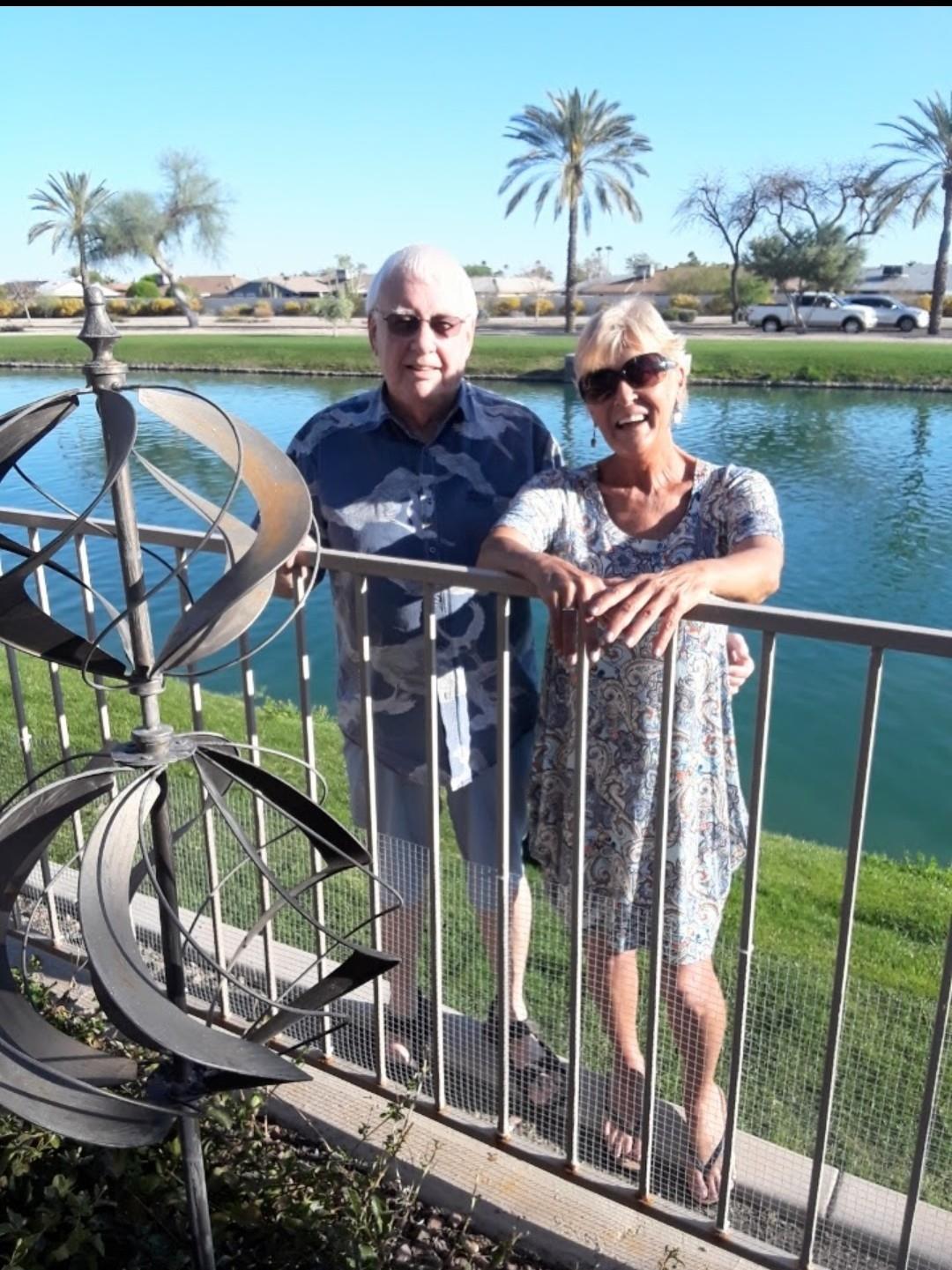 Two seniors smile while leaning against a railing by a serene lake surrounded by palm trees.