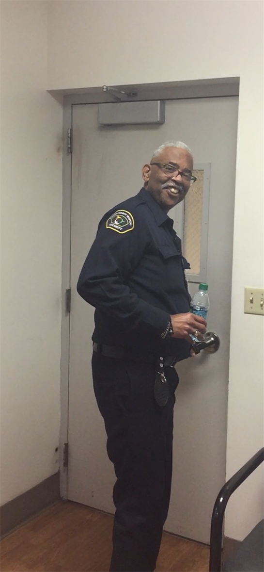A police officer stands by a door, smiling and holding a bottle of water during duty.