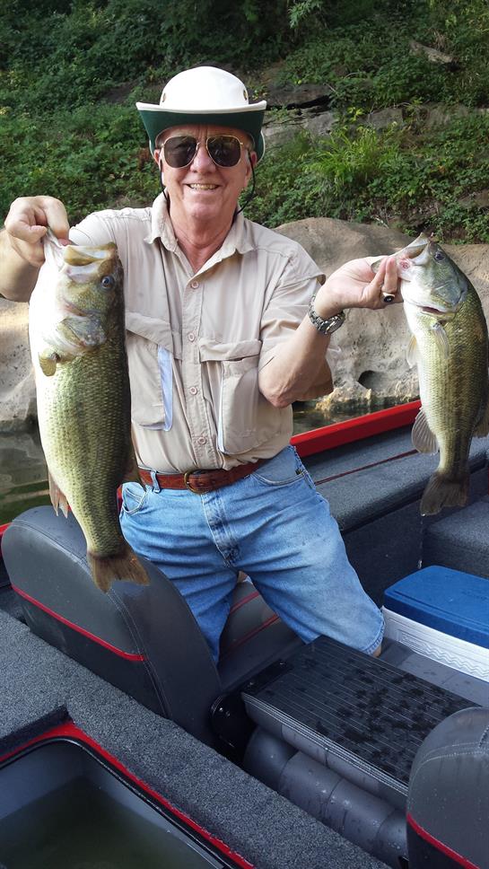 A man with sunglasses and a hat displays two large bass fish on a boat during a summer fishing trip.