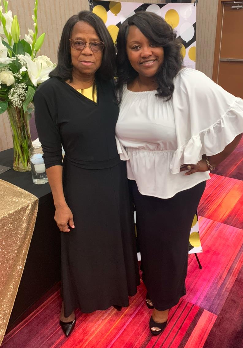 Two women stand side by side, smiling and dressed stylishly for a special occasion.