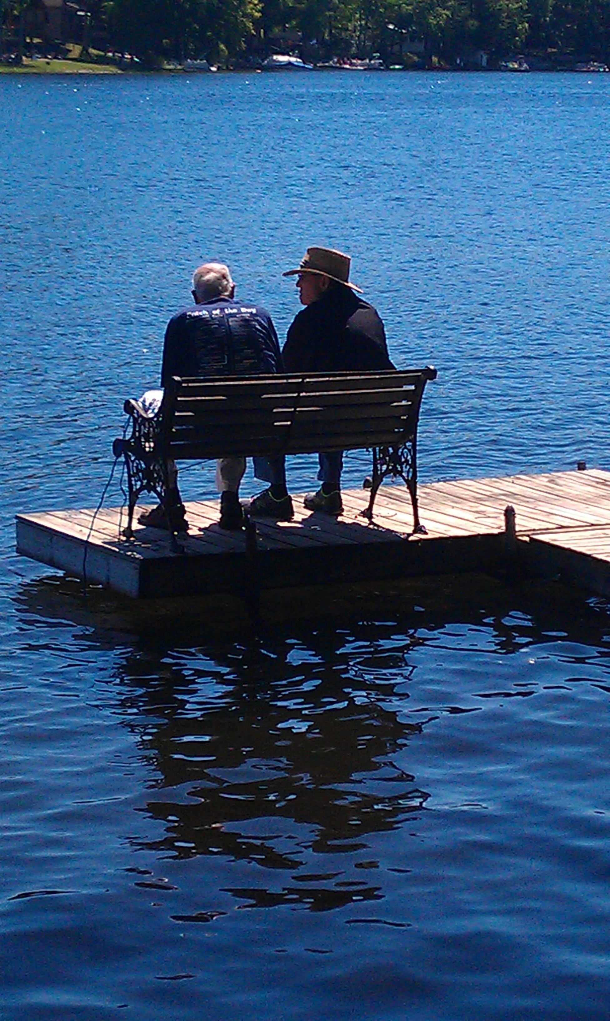Two individuals relax on a dock by a serene lake, enjoying conversation and fresh air.