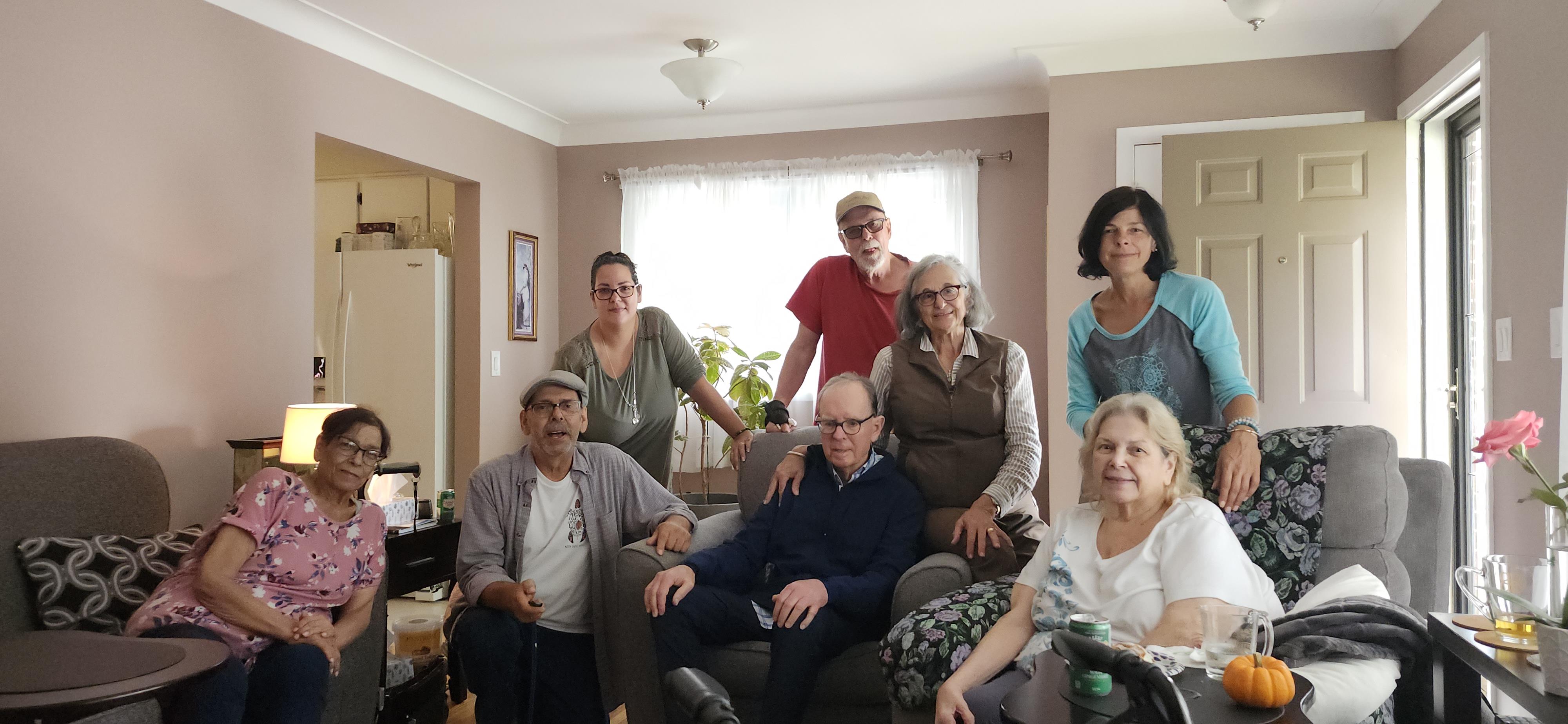 A group of eight adults, diverse in age and background, smiles together in a warm living room.