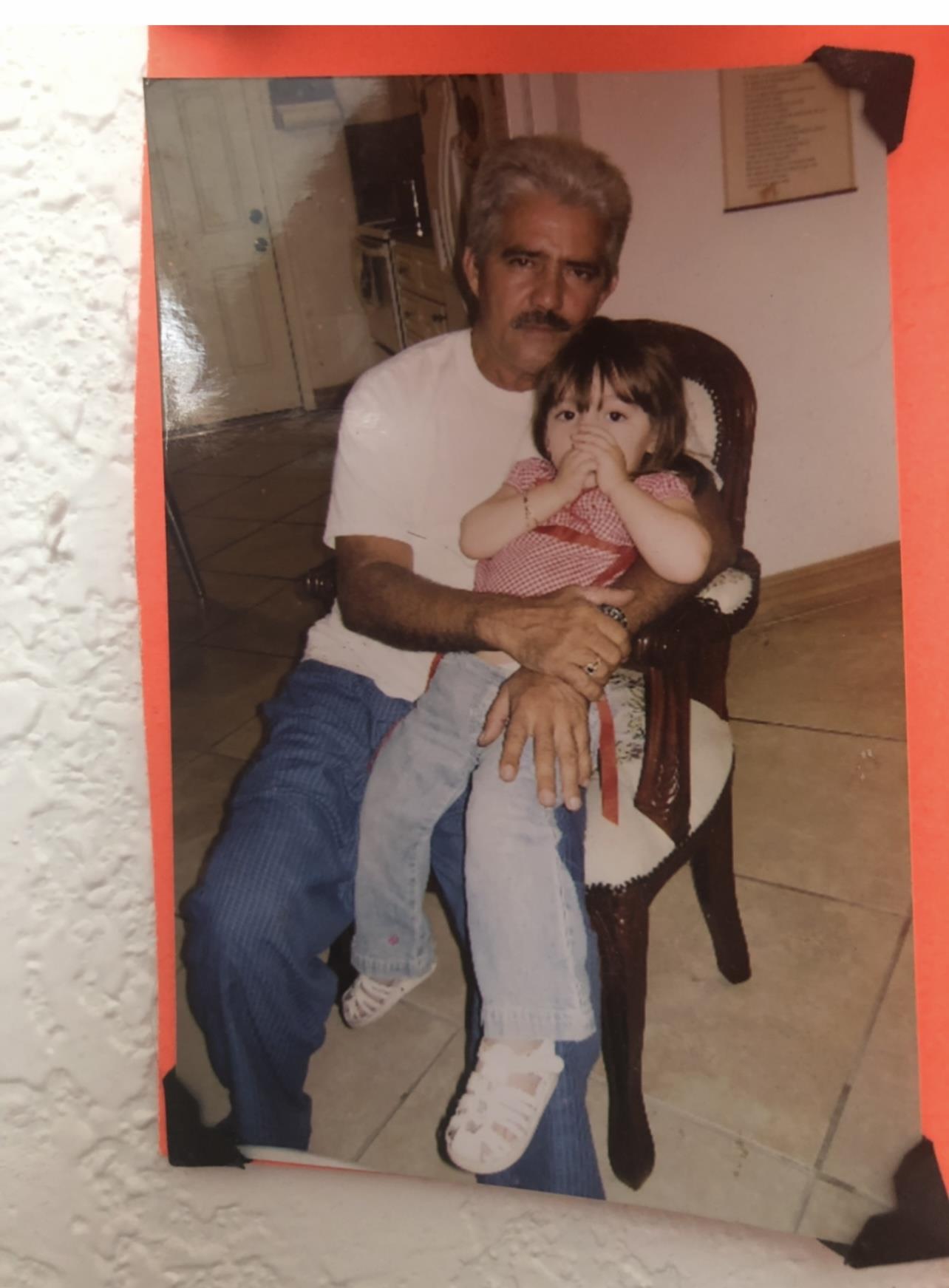 A loving grandfather sits in a chair, holding his granddaughter who looks curious and playful.