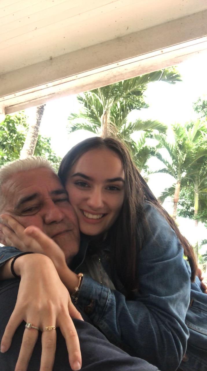 A daughter hugs her father while smiling, amidst lush palm trees and a bright atmosphere.